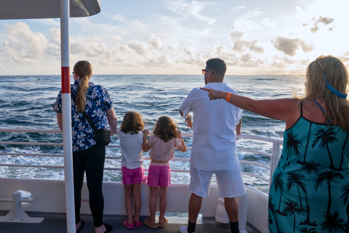 People on a boat looking at the ocean, woman in a blue dress pointing, with the sun setting.