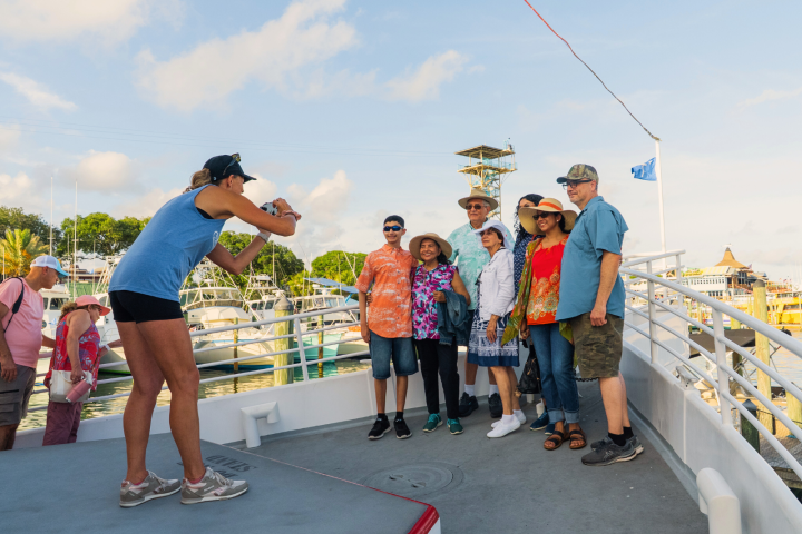 Person in blue tank top takes a photo of people posing on a boat with a marina in the background.