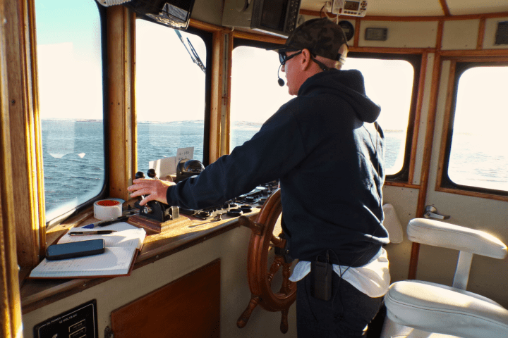 Person steering a boat inside a control room, wearing a cap and headset.