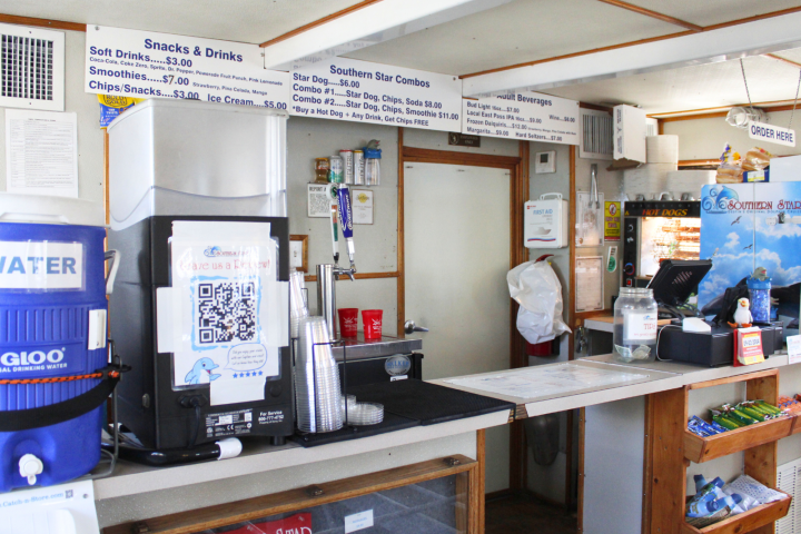 Concession stand with a drink dispenser, menu signs, QR code, and snacks on wooden shelves.