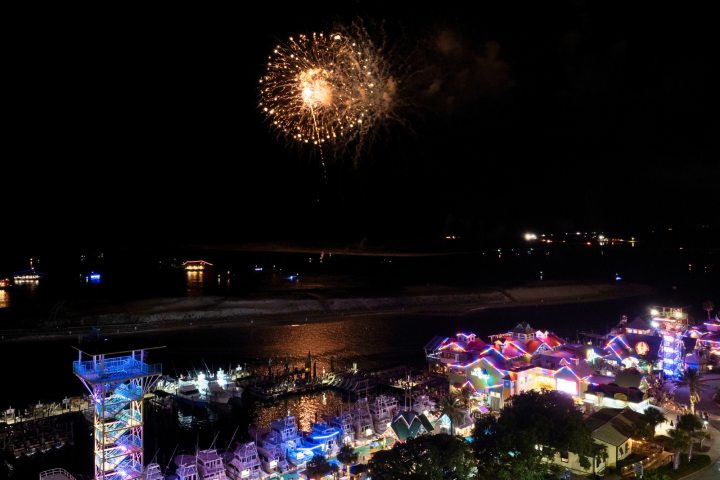 Night scene with fireworks over a brightly lit seaside town and marina.