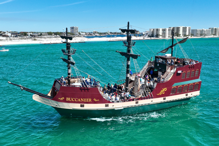 Pirate-themed ship 'Buccaneer' on turquoise water, with passengers on deck, distant shore and buildings visible.