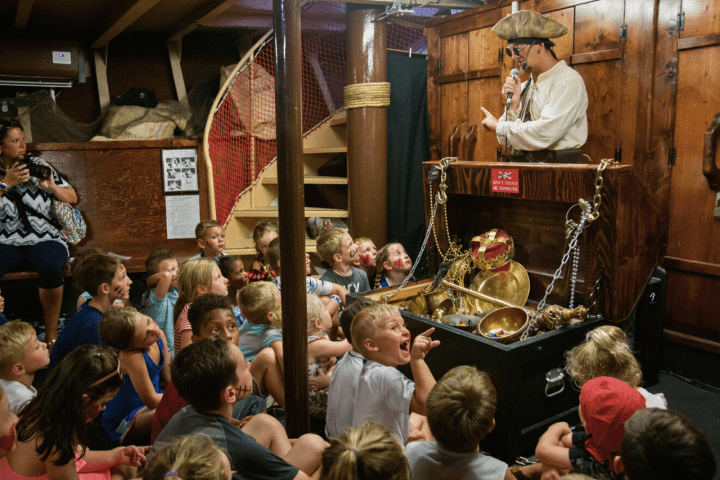 Pirate-themed storyteller captivates children with treasure chest in wooden ship interior.