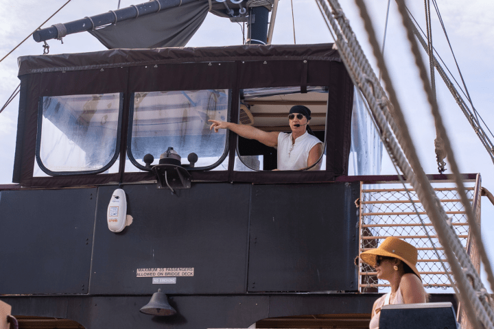 Person in pirate attire points from ship's bridge, another person in hat on deck.