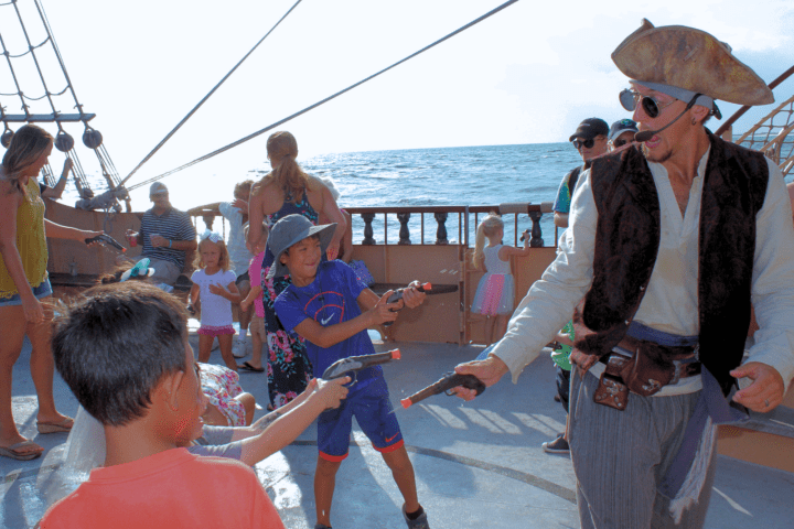 Kids with toy guns play with a pirate actor onboard a ship with ocean in the background.