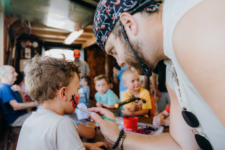 Man painting child's face with pirate theme in a lively indoor setting with kids watching.