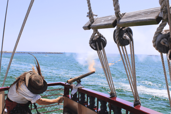 A person fires a cannon from a sailing ship on a sunny day with ocean view.