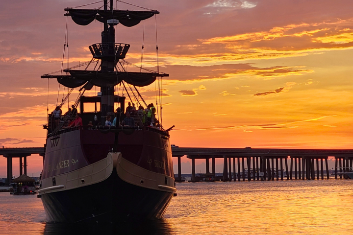 A pirate-themed ship sails into a vibrant orange sunset under a bridge.