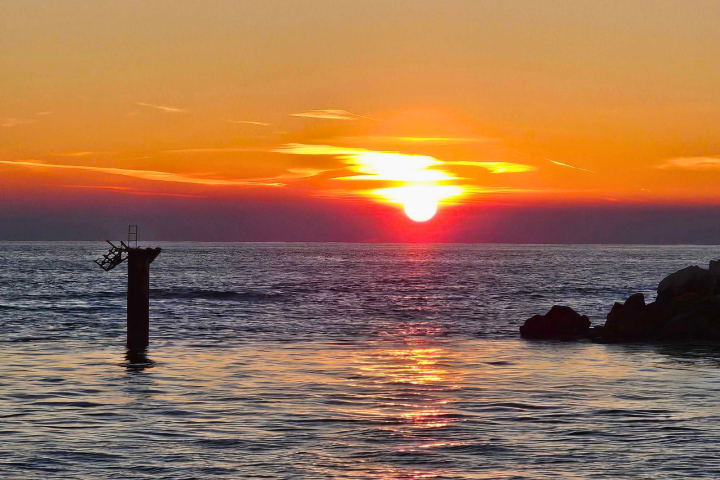 Sunset over ocean with a silhouetted pillar and rocks in the foreground.